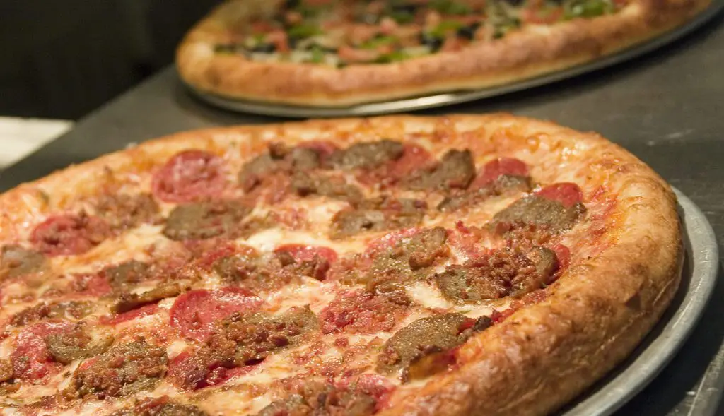 Close-up of a Manhattan meat lover's pizza with pepperoni, sliced meatballs, and melted cheese on a metal pan, with another pizza blurred in the background.