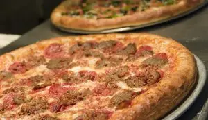 Close-up of a Manhattan meat lover's pizza with pepperoni, sliced meatballs, and melted cheese on a metal pan, with another pizza blurred in the background.
