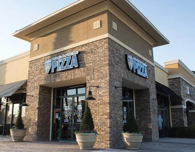 Corner view of the NYPD Pizza Hunter's Creek restaurant with a brick exterior, tall entryway, and large potted plants near the front doors.