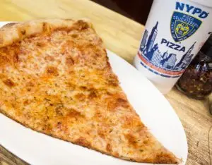 Large slice of cheese pizza on a white plate next to an NYPD Pizza branded cup.