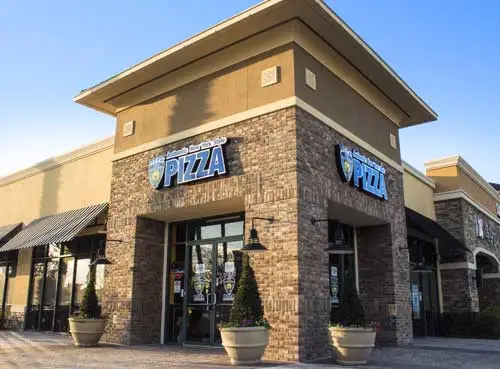 Corner view of the NYPD Pizza Hunter's Creek restaurant with a brick exterior, tall entryway, and large potted plants near the front doors.