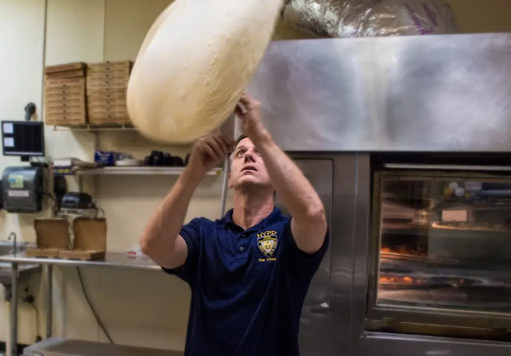 NYPD Pizza founder and CEO Paul Russo tosses a large sheet of pizza dough into the air inside a commercial kitchen.