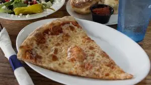 Large slice of New York–style cheese pizza on a white plate, served at a restaurant table.